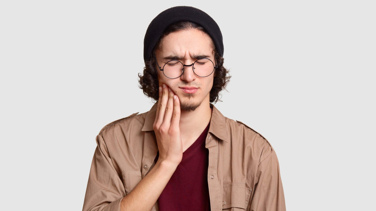 Photo of stressful youngster with small beard keeps hand on cheek, suffers from toothache, keeps eyes shut, dressed in stylish clothes, big round spectacles, models against white studio wall